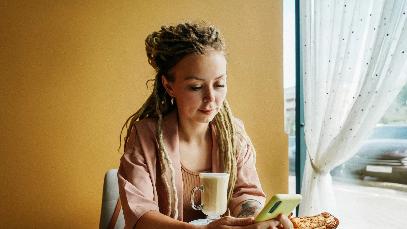 Jeune femme lisant ses e-mails sur son téléphone dans un café, un latte à la main : email marketing et scénarios automatiques