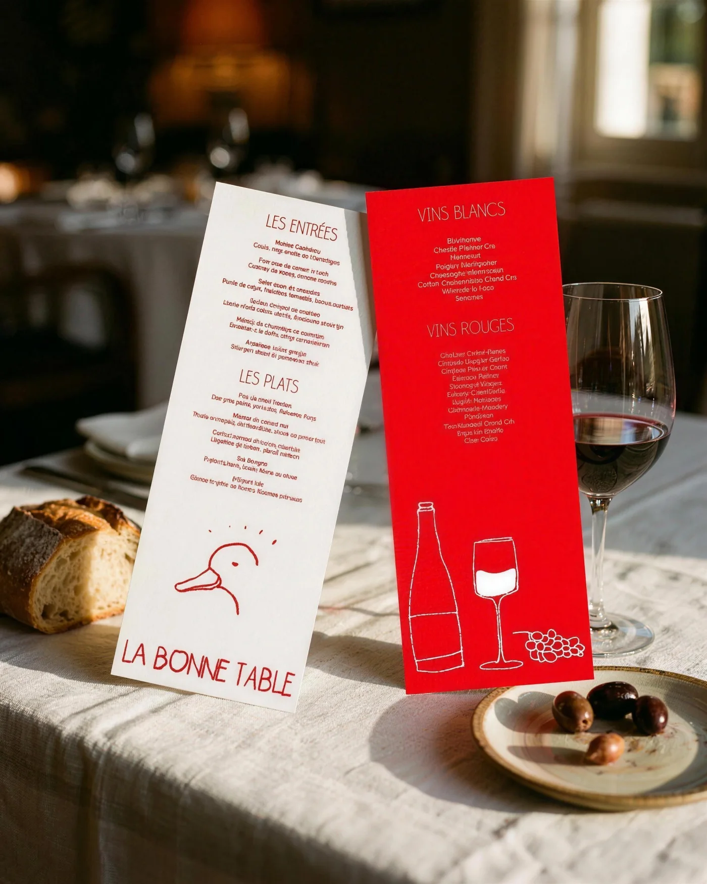 The red and white La Bonne Table menus on a linen tablecloth with bread, olives and a glass of red wine