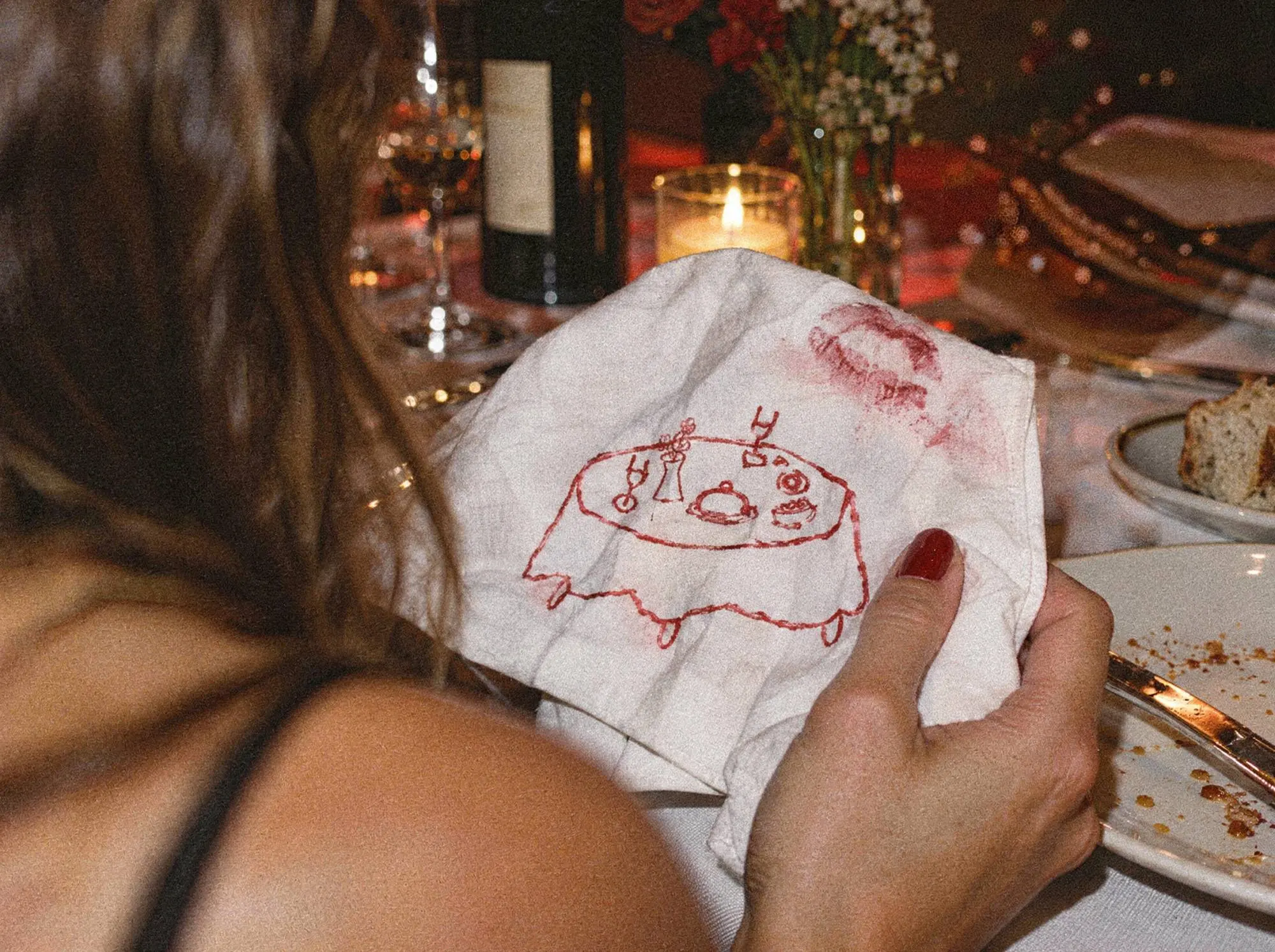 A guest holds her napkin with the red table drawing and a lipstick mark, candles and wine on the table