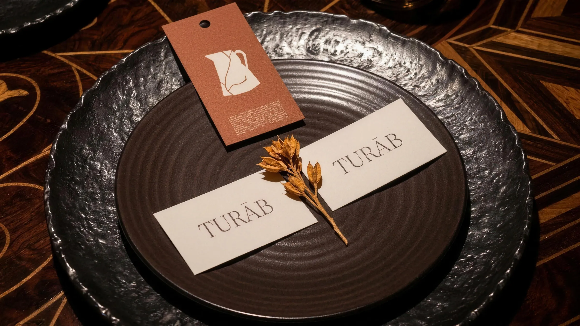 Turāb hang tag with the cracked jug and two Turāb cards with a dried flower on a dark stoneware plate in a hammered metal tray